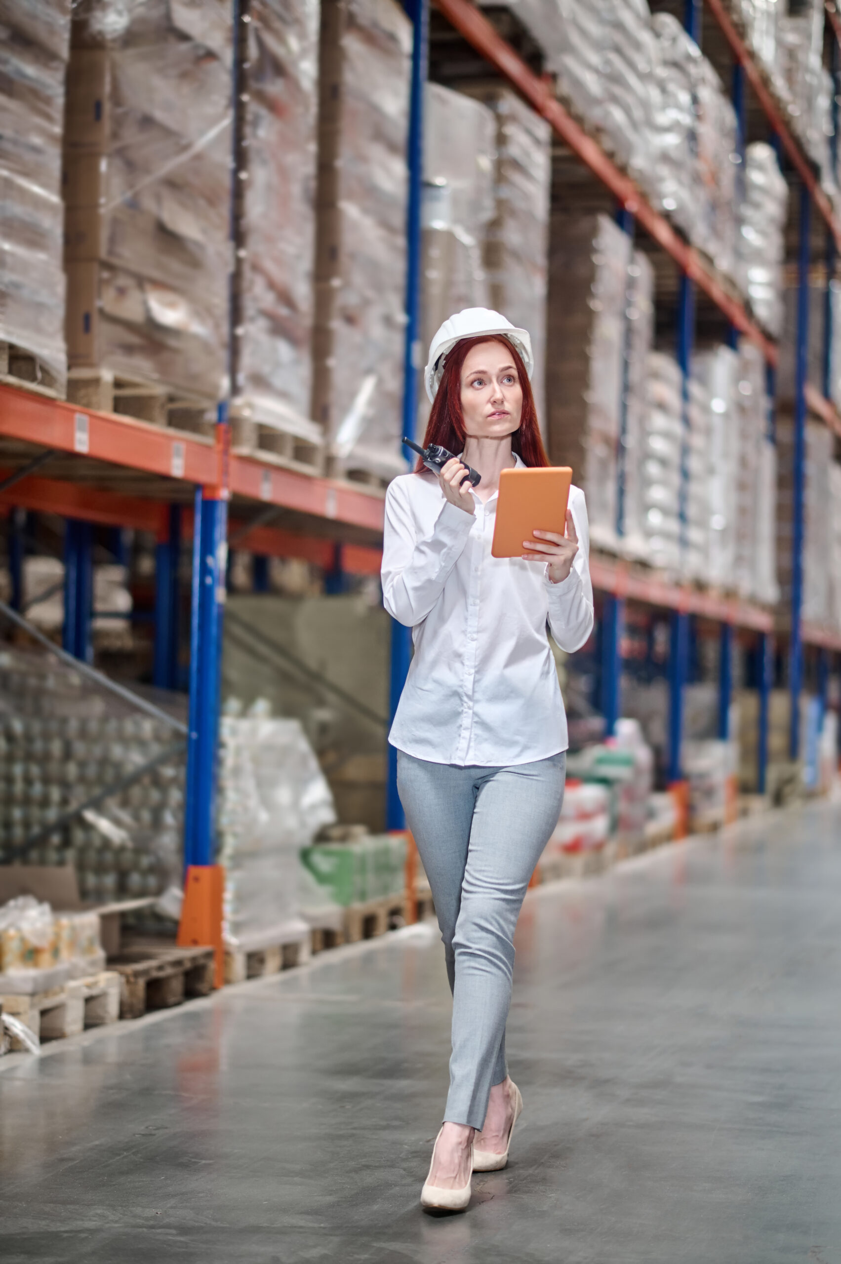 Check, warehouse. Attentive elegant woman with long red hair in safety helmet with tablet and walkie-talkie looking up and aside checking at warehouse